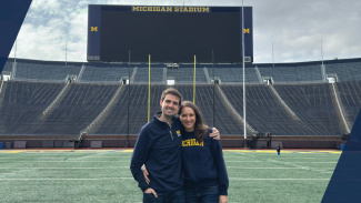 A couple smiles, standing together on the field of the Big House with the jumbotron screen and goalpoast behind them.