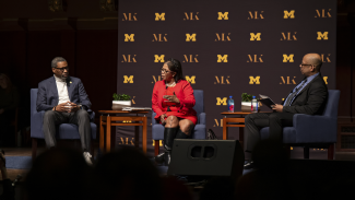 Three speakers have a panel discussion on stage with University of Michigan branding in the background.