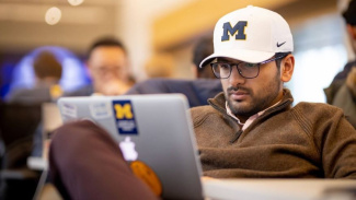 Student wearing a University of Michigan hat works on a laptop with Michigan stickers.