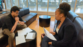 Two people sit on couches discussing documents spread out on a table.