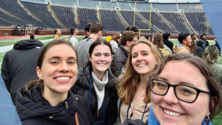 Four people smiling on a football field with stadium seats in the background.