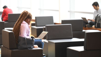 Photo of a woman sitting in a chair looking at her computer. She is wearing a pink sweatshirt and has long braided brown hair. There are two students in the background that are also looking at computers but their faces are blurred out. 
