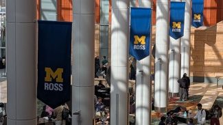 Photo of the interior of the Ross School of Business Winter Garden. There are multiple maize and blue Michigan Ross banners hung on the poles. There are students sitting at tables as well.