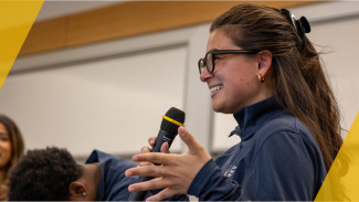 Smiling person wearing glasses speaks into a microphone during an indoor event or presentation.