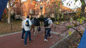 Four people with backpacks walk on a brick path toward a university building in autumn.