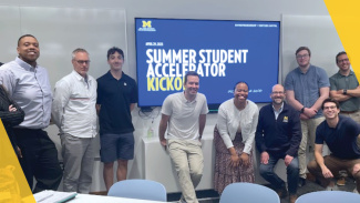 Group of people standing and smiling in front of a screen displaying "Summer Student Accelerator Kickoff."