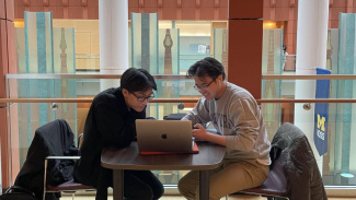 Two Michigan Ross students sitting at a table with a laptop computer between them. Both students are male and have short dark hair and both are wearing glasses. There are white pillars in the background with Michigan Ross flags on them.