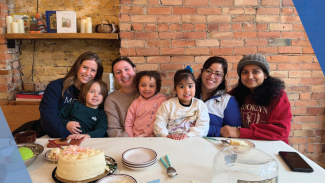 Six people sit at a table with a cake, smiling in front of a brick wall.