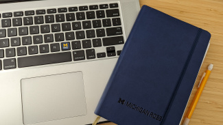 Photo of a computer keyboard next to a Michigan Ross blue notebook. There is a yellow pencil next to the notebook on the table. 