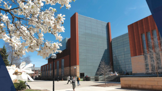 Modern university building with glass and orange paneling, framed by white cherry blossoms in the foreground.