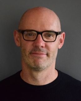 Headshot of a bald man with stubble wearing black-framed glasses against a gray background.