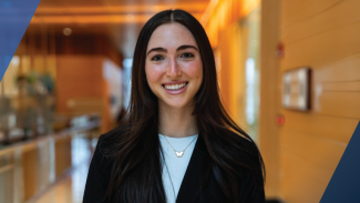 Smiling person with long dark hair standing indoors in a modern building.