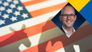 Man in glasses and dark blazer beside a Democratic donkey logo over an American flag background.