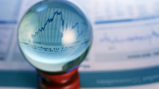 A clear glass sphere sits in front of financial charts, enlarging a section of a blue line graph. The background is intentionally blurred, emphasizing the magnified market trend and suggesting analysis or economic uncertainty.