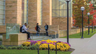 Students sit and chat on benches outside a stone campus building surrounded by autumn flowers and trees.
