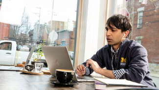 Man sitting at a table looking at his laptop. He is wearing a Michigan Ross sweatshirt and he has dark hair. There is a coffee cup next to his computer and there is a white truck outside the window he is sitting near. 