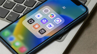 A smartphone resting on a laptop keyboard displays a folder labeled "AI" containing icons for several popular AI assistant apps, including ChatGPT, Gemini, Copilot, Meta AI, Claude, Grok, DeepSeek, Perplexity, and others.