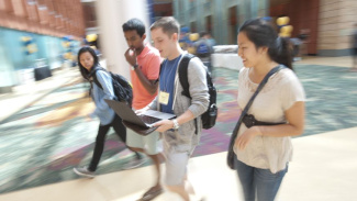 Four students walking through the Michigan Ross Winter Garden. There are two males and two females, they are all wearing backpacks and one is carrying a laptop computer. 