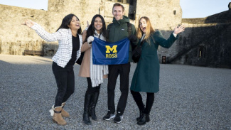 Four students, three females, one male, standing together holding a maize and blue Michigan Ross flag. The two students on the end have their arms up in the air in excitement. There is a castle in the background. 