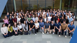 Large group of people posing together outside a modern glass building.