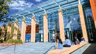 Modern glass-fronted university building with students sitting on outdoor steps on a sunny day.