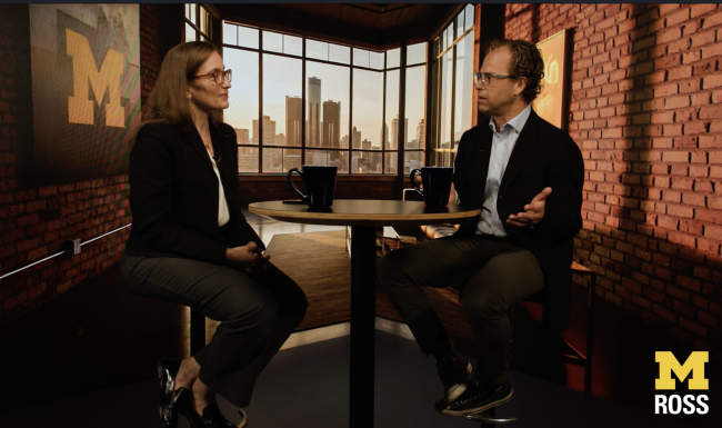 Justin Wolfers and Stacey Vanek Smith sit across from each other at a round table in a studio with a city skyline backdrop and large windows. Both are dressed in business attire, engaged in conversation with coffee mugs on the table. The scene features warm lighting, brick walls, and a yellow “M ROSS” logo in the lower right corner.