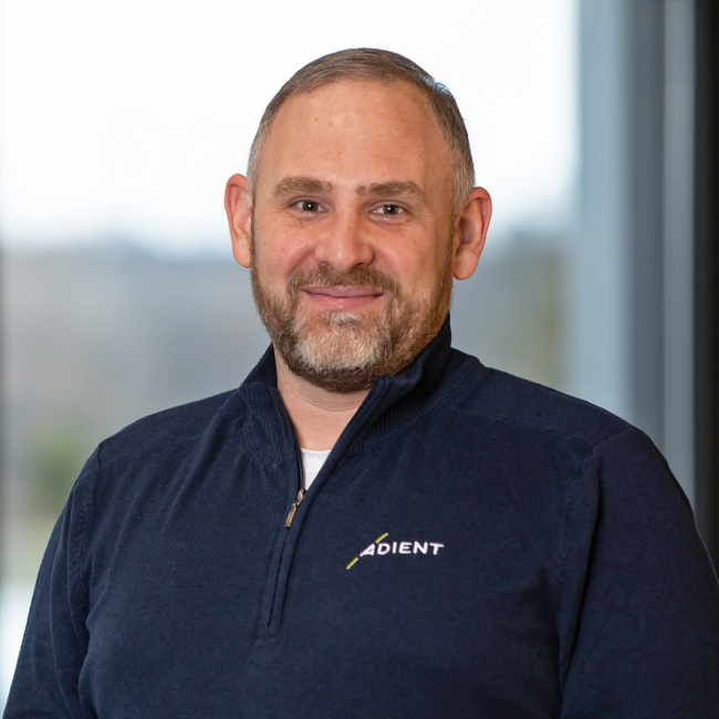 Smiling person wearing a dark shirt with the Adient logo, standing indoors.