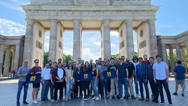 A group of people pose in front of the Brandenburg Gate, some holding a University of Michigan flag.