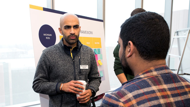 A person speaks with another attendee in front of a presentation board at a professional event.