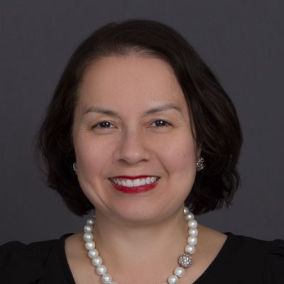 Woman with short dark hair, red lipstick, pearl necklace, and black top smiling against gray background.