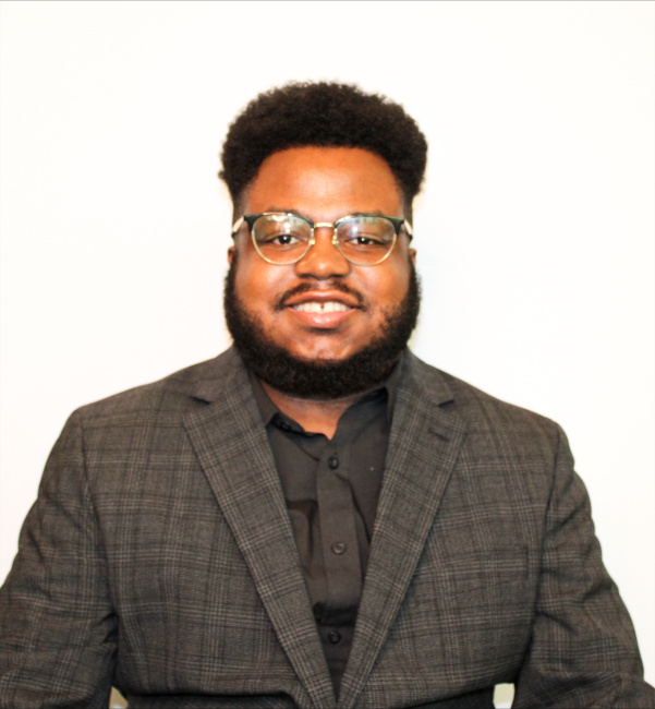 Rasul Rahman wearing glasses and a dark plaid suit jacket smiles in front of a plain white background.