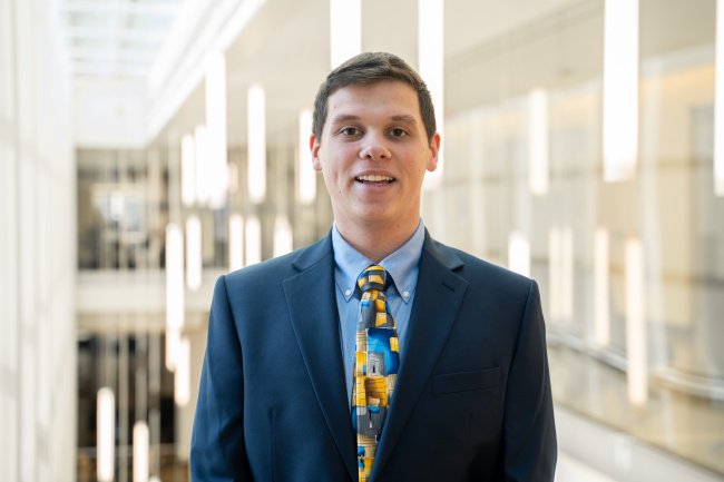 Man in a dark suit and patterned tie smiling in a bright modern building interior.