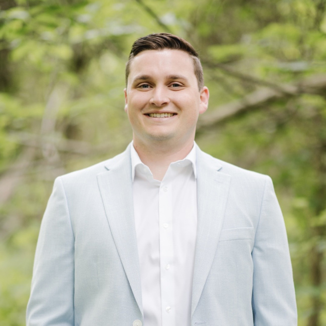 Man in a light suit jacket smiling outdoors with greenery in the background.