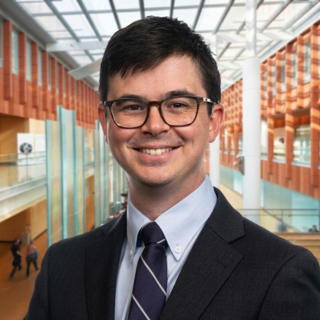 Smiling man in a suit and glasses standing in a modern office atrium.