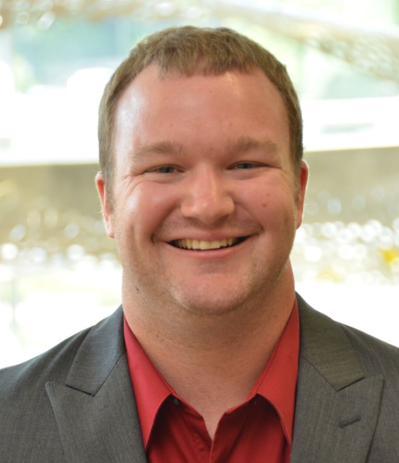 Smiling person wearing a red shirt and gray blazer.