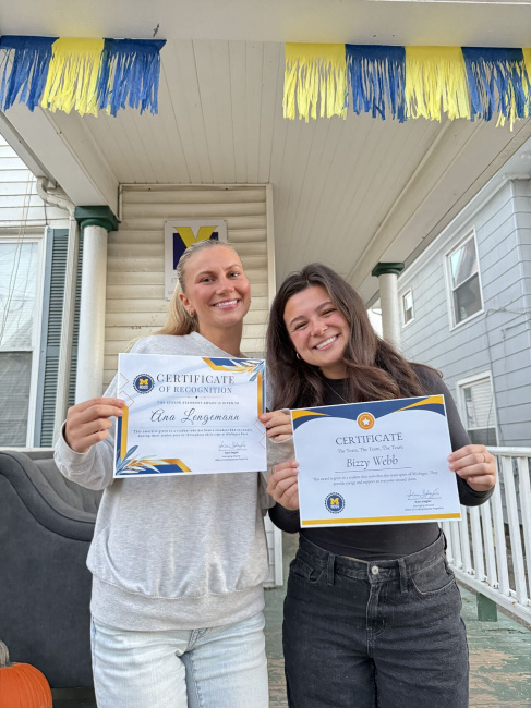 Two women pose smiling with certificates on the porch of a house with a maize and blue banner.