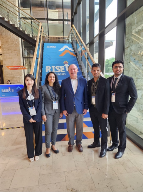 Five people in business attire stand in front of a staircase with a "RISE" conference banner.