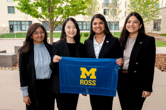 Four women in business attire hold a University of Michigan Ross banner outdoors.
