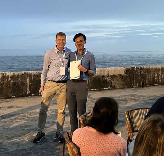 Two people stand by the ocean at sunset, one holding a certificate during an award presentation.