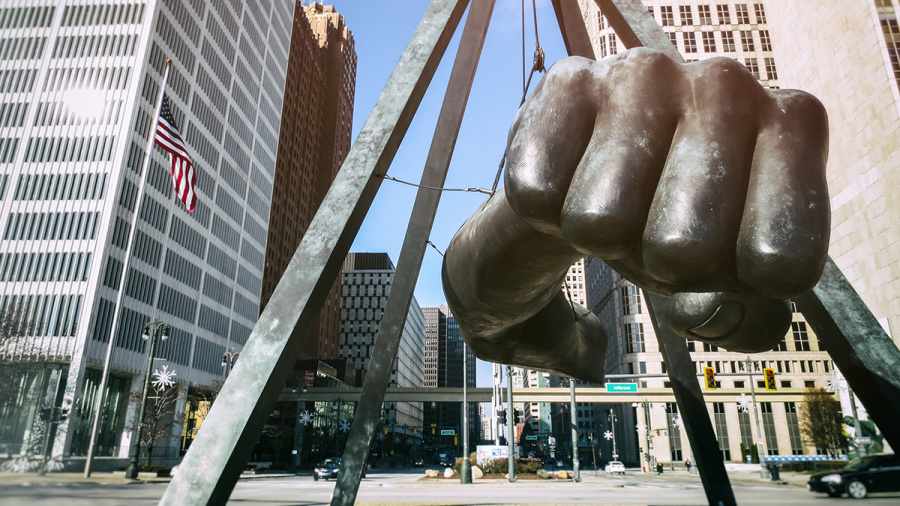 Large bronze fist sculpture suspended above a downtown city street with office buildings in the background.