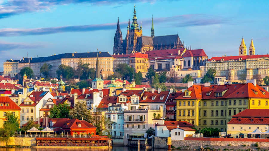 A panoramic view of Prague, showing colorful buildings in the foreground and the historic Prague Castle with its Gothic architecture in the background.