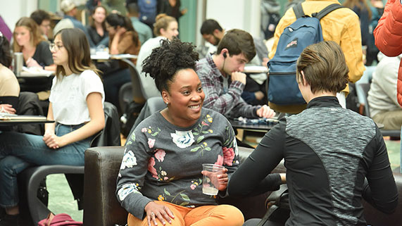 A group of people socializing and studying in a bustling indoor space, featuring varied clothing styles and multiple tables.