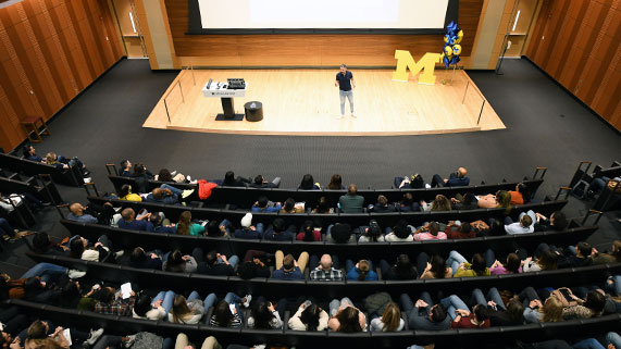 A speaker presents to a packed lecture hall, while students take notes in a modern auditorium setting.