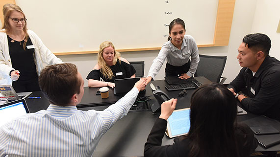 A group of professional students engage in a collaborative meeting, with one person extending a handshake across the table. 