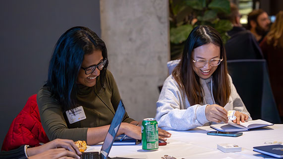 Two people engaged in a collaborative work session at a table, with laptops, a notebook, snacks, and a green drink nearby.