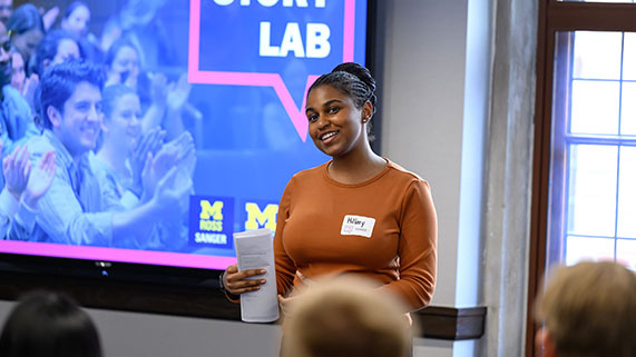 A presenter stands in front of a screen at an event, holding a paper and engaging with the audience.