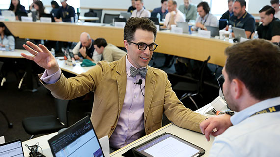 professor gestures animatedly during a classroom discussion, while students engage at their desks, using laptops and tablets.