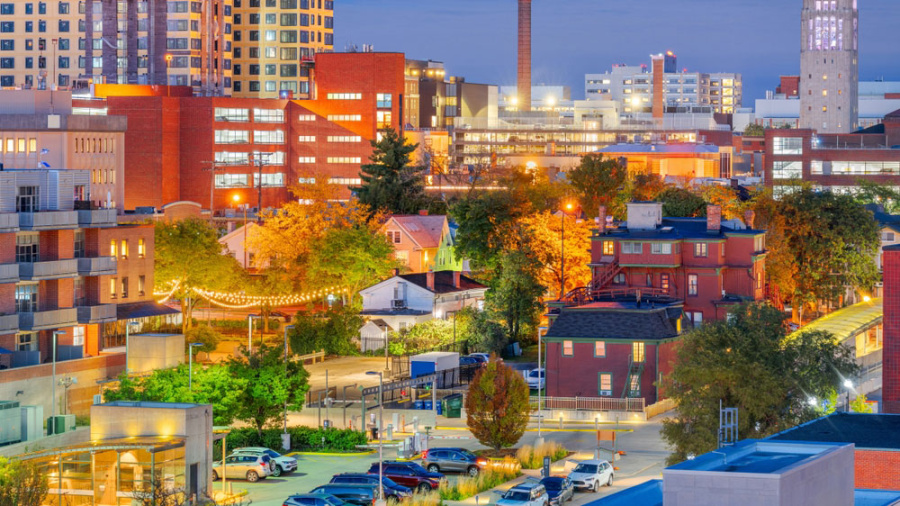 A vibrant cityscape at dusk, featuring illuminated buildings and apartments. Trees with autumn foliage surround cozy houses and a well-lit street.