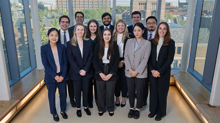 Twelve professionally dressed people stand together in a modern building with large windows and a city view.