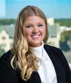 Person with long blonde hair in business attire smiling in an office with a city view.